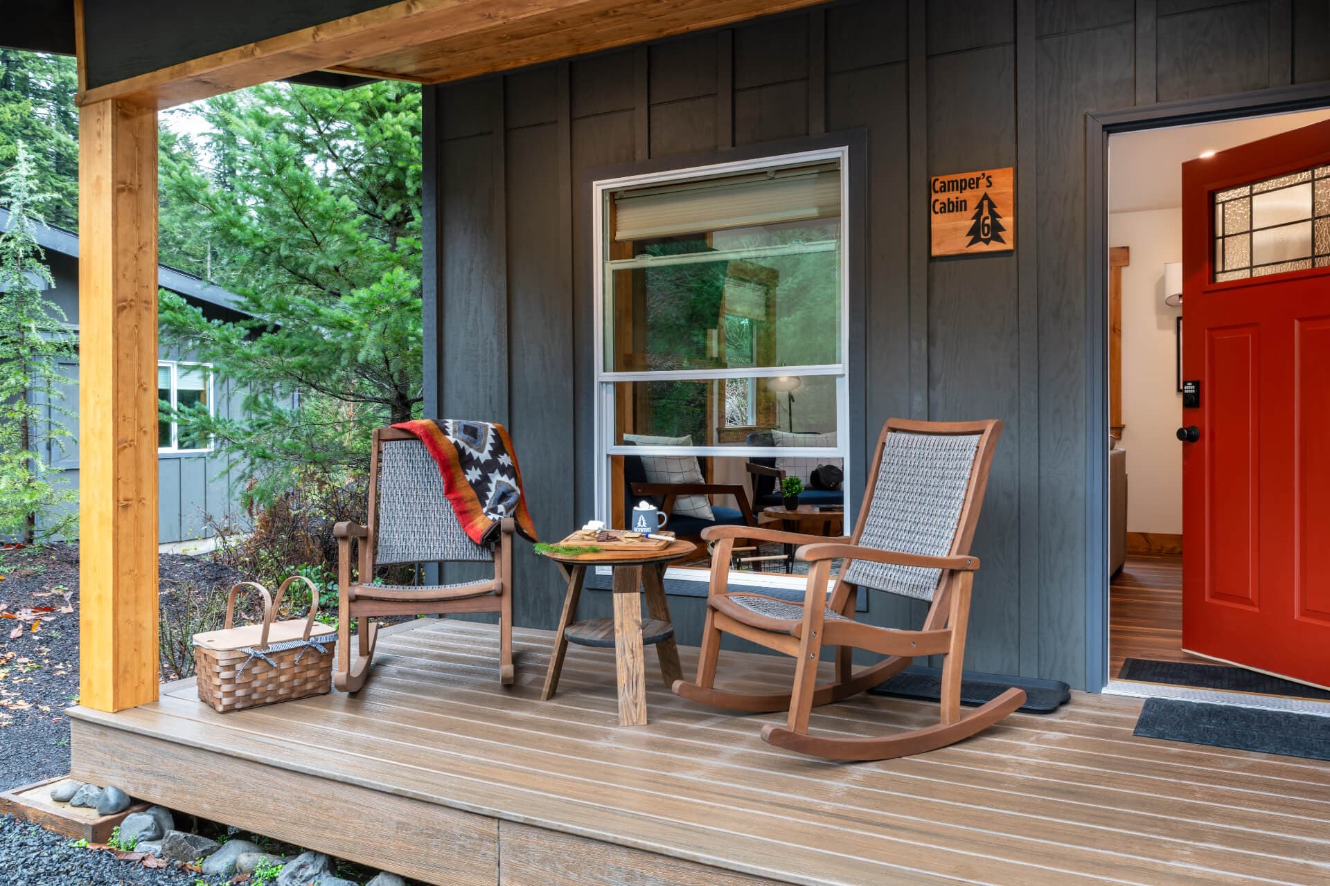 A cozy porch features two rocking chairs, a small table, and a basket, with a welcoming red door and greenery in the background.