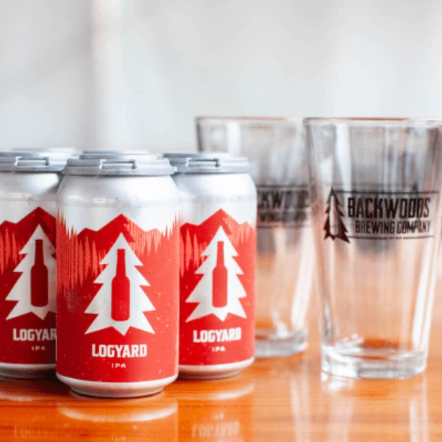Four cans of Logyard IPA beside two Backwoods Brewing Company glasses on a wooden surface.