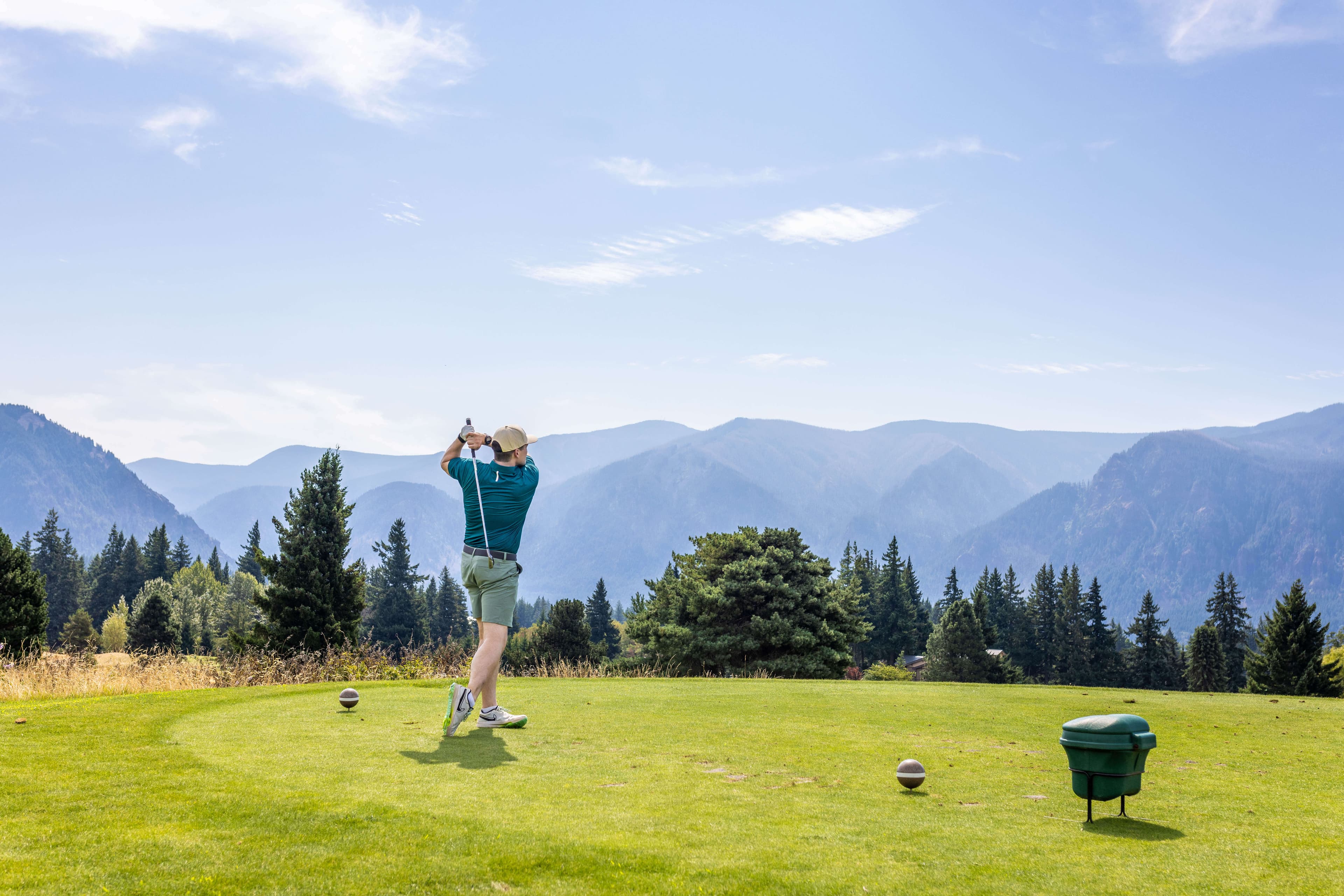 A golfer swings on a tee with mountains in the background.
