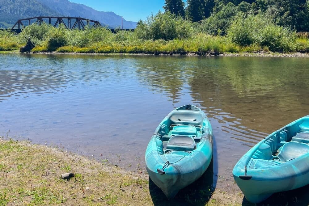 Two blue kayaks are moored along a calm riverbank with a train bridge in the background.