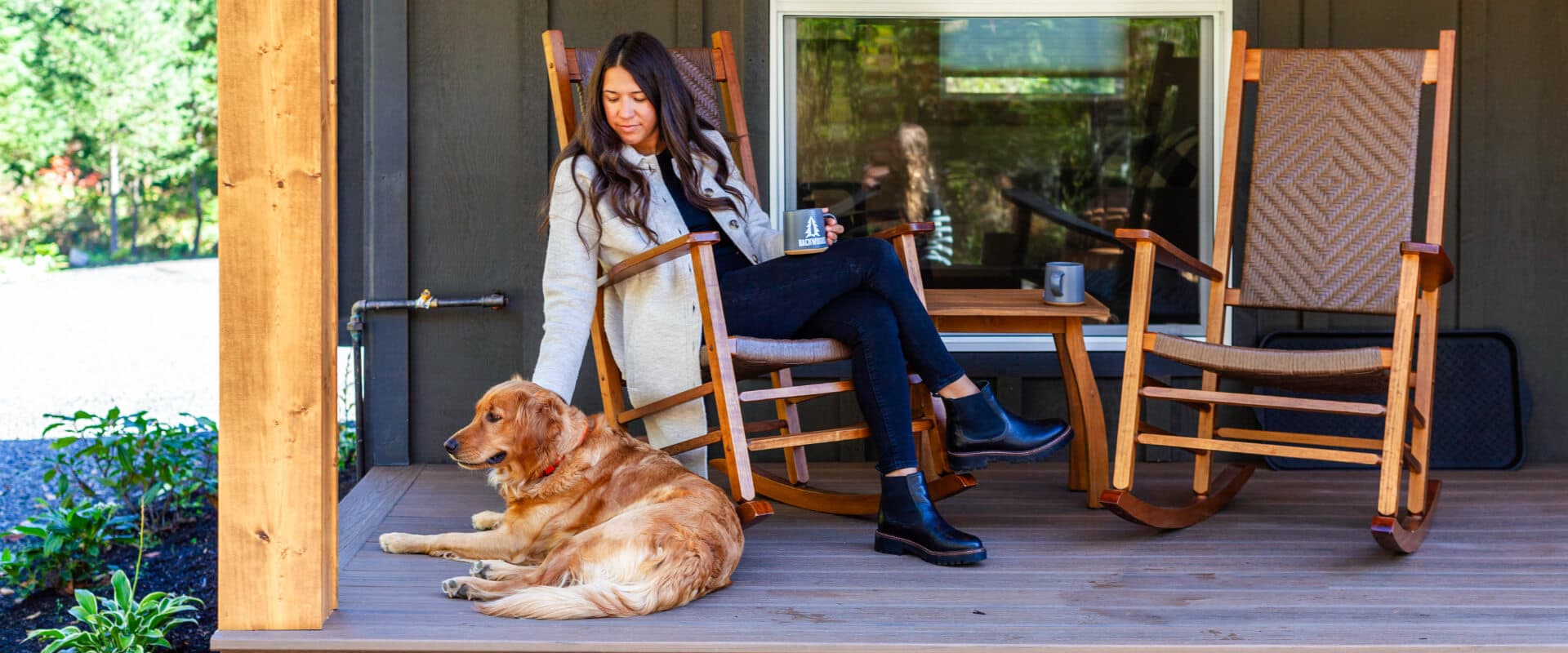 A woman sits on a porch beside a golden retriever, enjoying a coffee in a cozy outdoor setting.