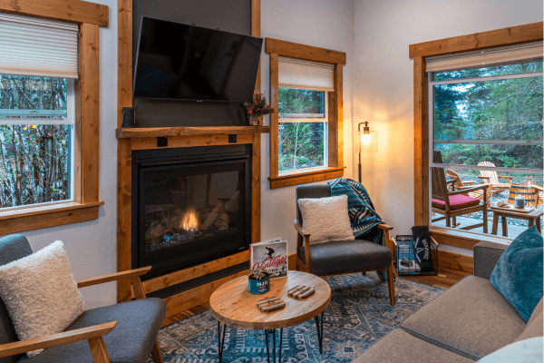 Cozy cabin sitting area with warm wood walls, a glowing fireplace, comfy chairs, and big windows looking out to the trees.