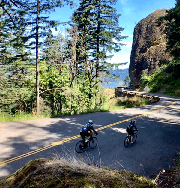 Two cyclists ride along a winding road surrounded by trees and rocky cliffs.