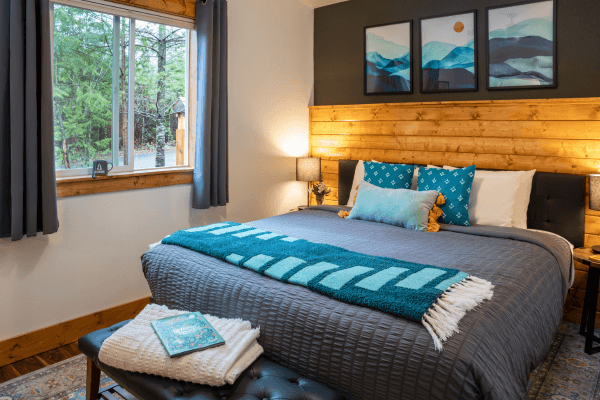 Warm cabin bedroom with a neatly made bed, wood accents, soft lighting, and peaceful forest views through the window.