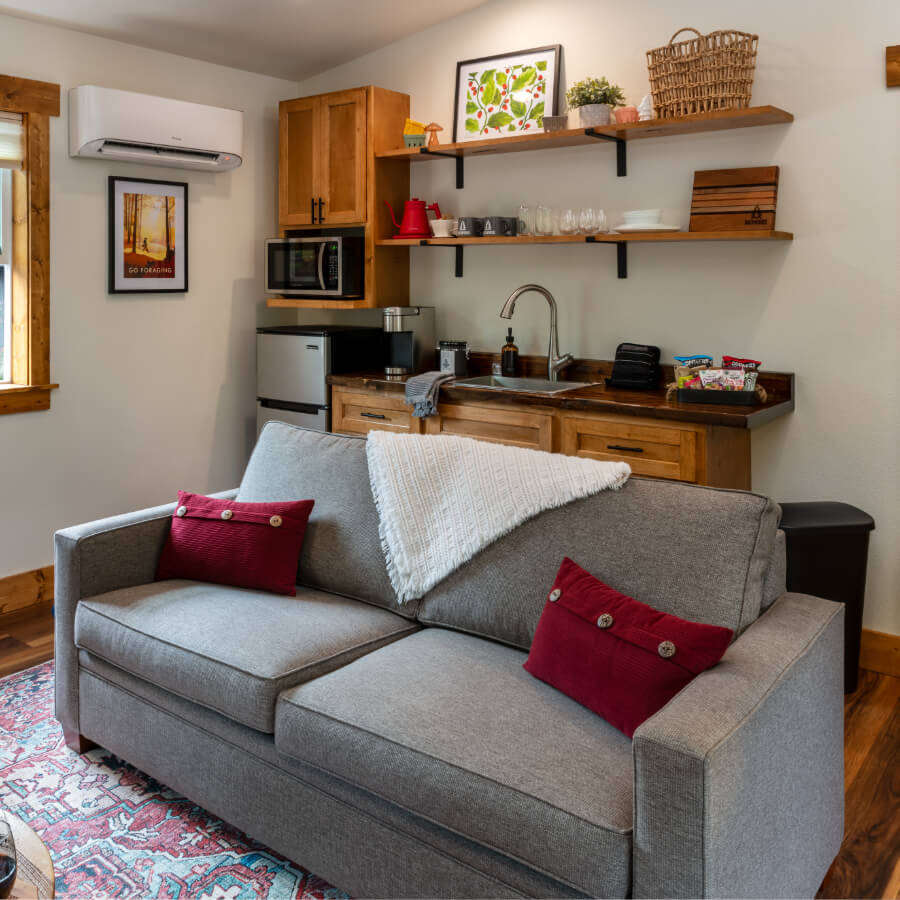Cozy living space featuring a gray couch with red cushions, a wall-mounted air conditioner, and a kitchenette with wooden shelves.