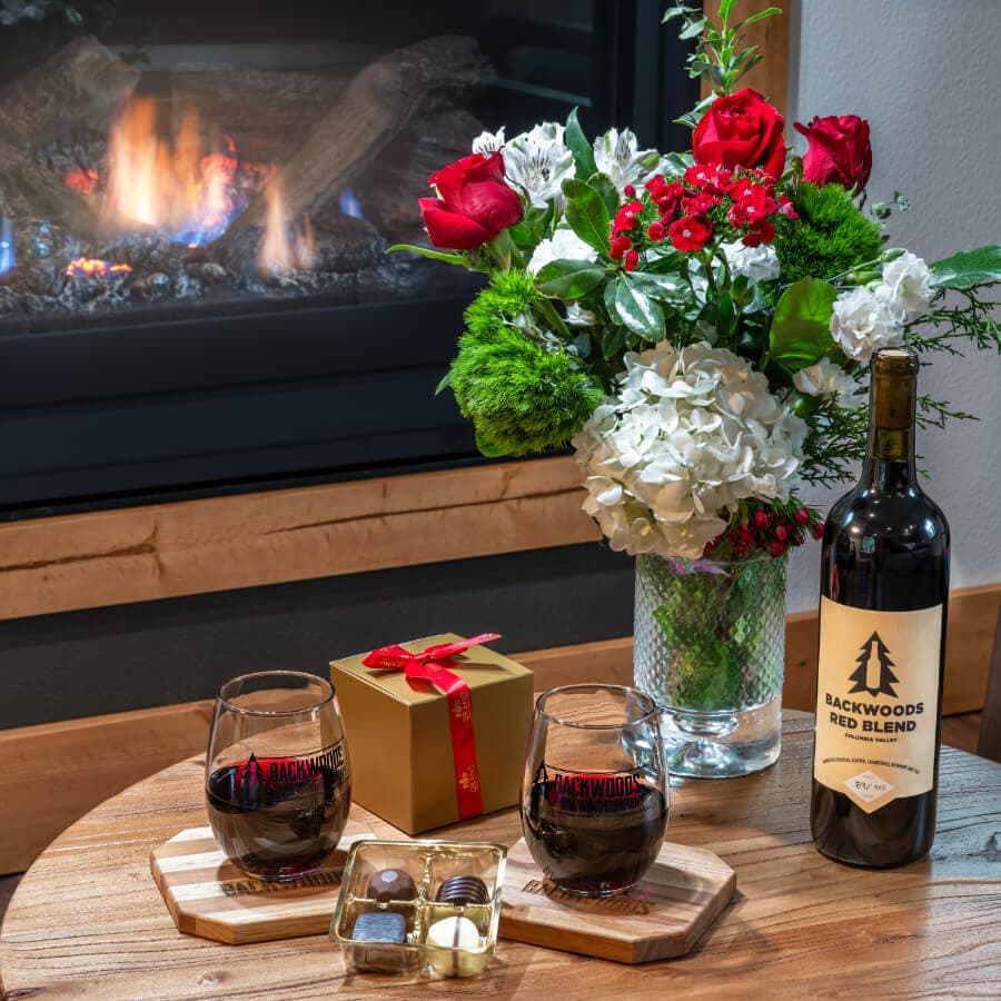 A cozy scene featuring a fireplace, a bottle of red wine, two glasses, chocolates, a gift box, and a floral arrangement.