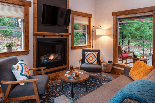 Cozy cabin sitting area with warm wood walls, a glowing fireplace, comfy chairs, and big windows looking out to the trees.