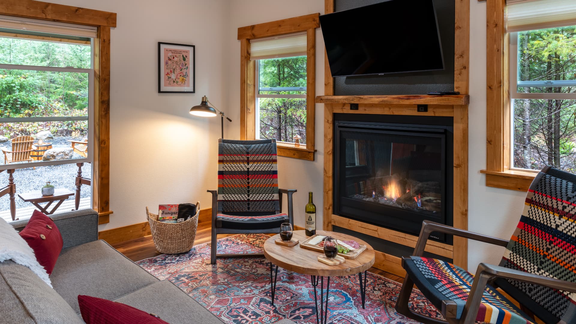 Cozy living room featuring two patterned chairs, a fireplace, a small table with drinks, and large windows overlooking a natural landscape.