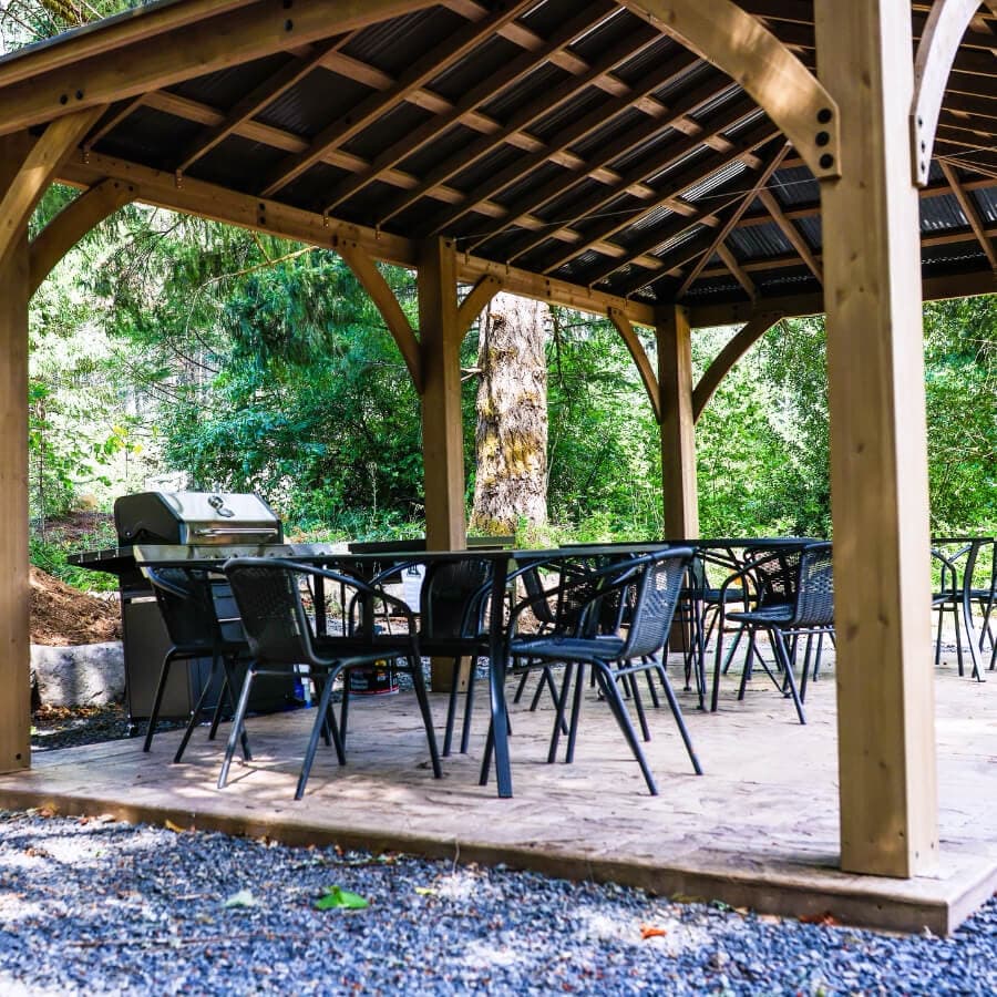 A covered outdoor seating area with several black chairs and a grill surrounded by greenery.