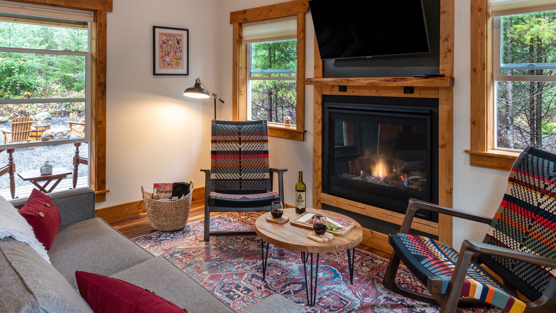 Cozy living room with a fireplace, two patterned chairs, a sofa, and large windows overlooking a forest.