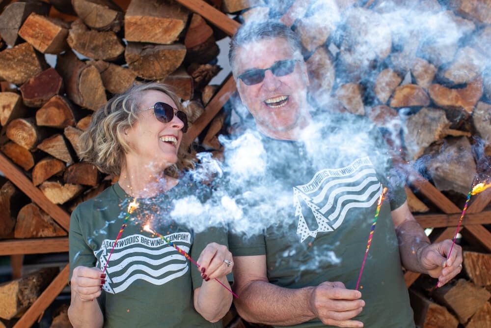A smiling couple holds sparklers against a backdrop of stacked firewood.