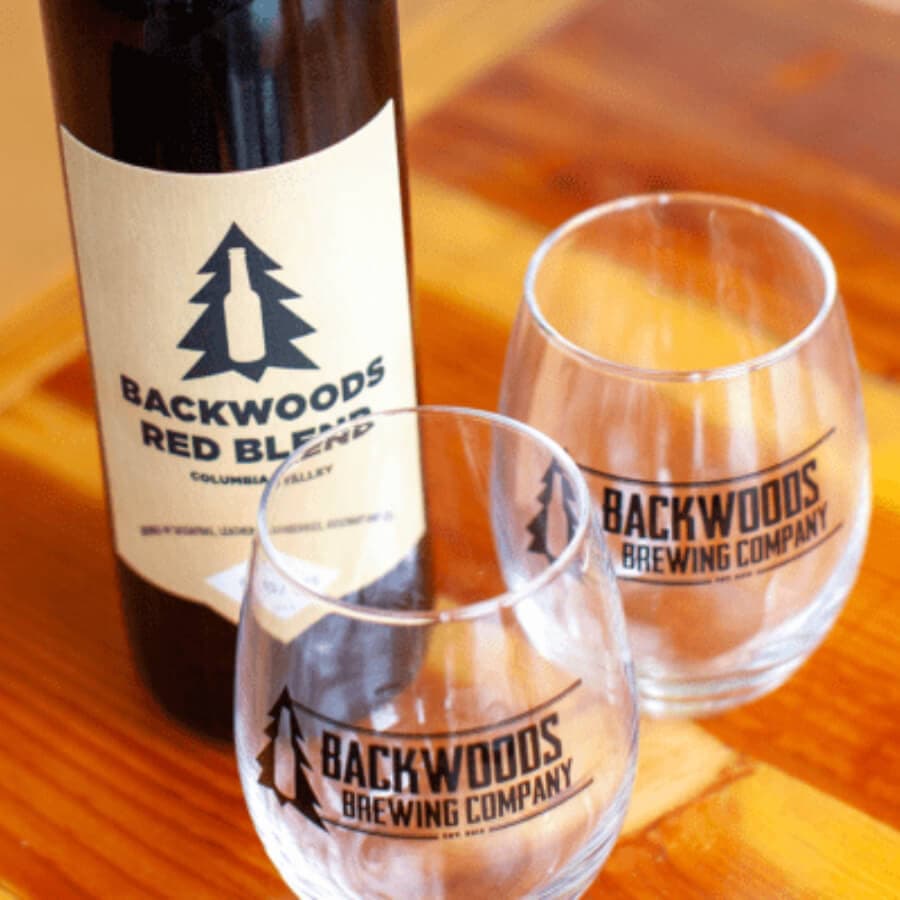 A bottle of Backwoods Red Blend wine alongside two logo-etched glasses on a wooden surface.