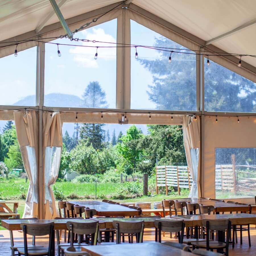 Interior of a spacious tent with wooden tables, overlooking a green outdoor landscape through large windows.