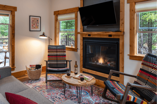 Cozy cabin sitting area with warm wood walls, a glowing fireplace, comfy chairs, and big windows looking out to the trees.