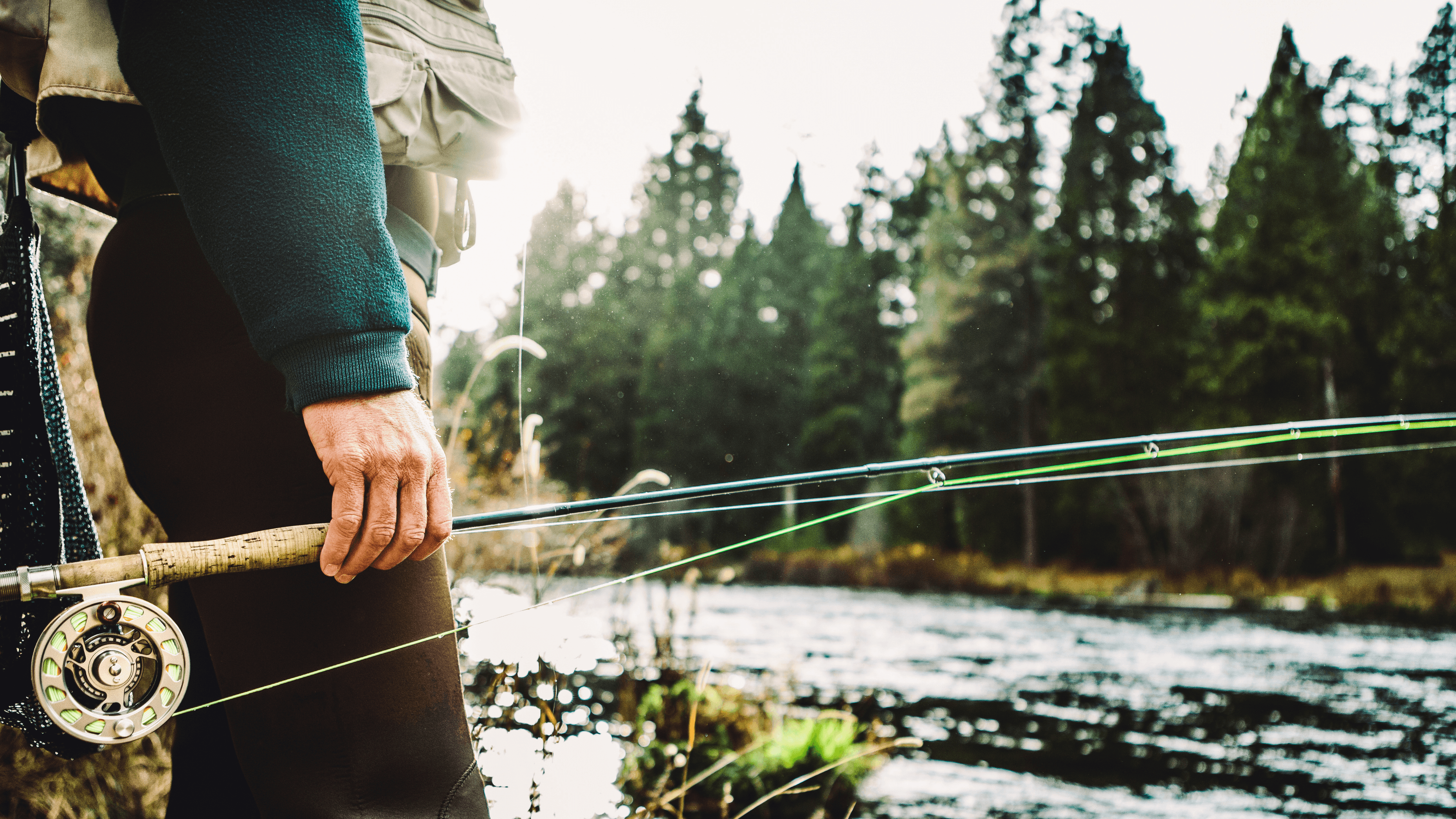 A person stands by a river, holding a fishing rod with vibrant lines, surrounded by lush evergreen trees.