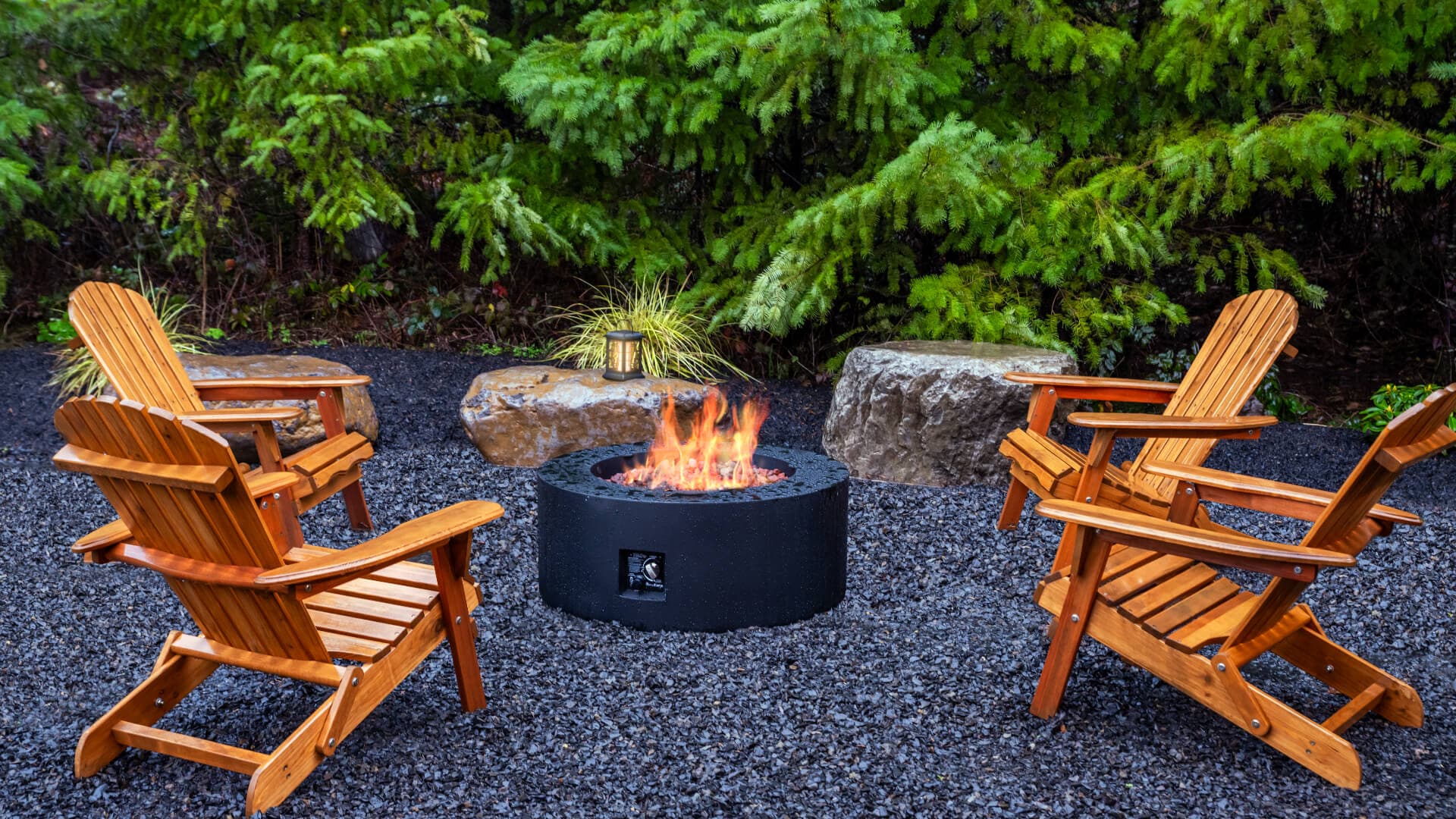 A cozy outdoor seating area with wooden chairs surrounding a fire pit, set against a backdrop of lush greenery.
