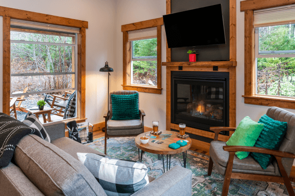 Cozy cabin sitting area with warm wood walls, a glowing fireplace, comfy chairs, and big windows looking out to the trees.