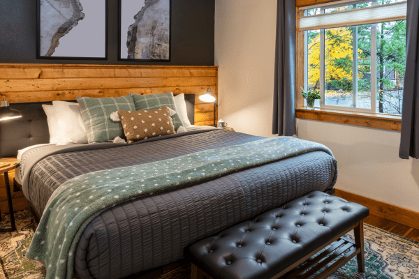 Cozy cabin bedroom with a neatly made bed, warm wood accents, soft lighting, and peaceful forest views through the window.