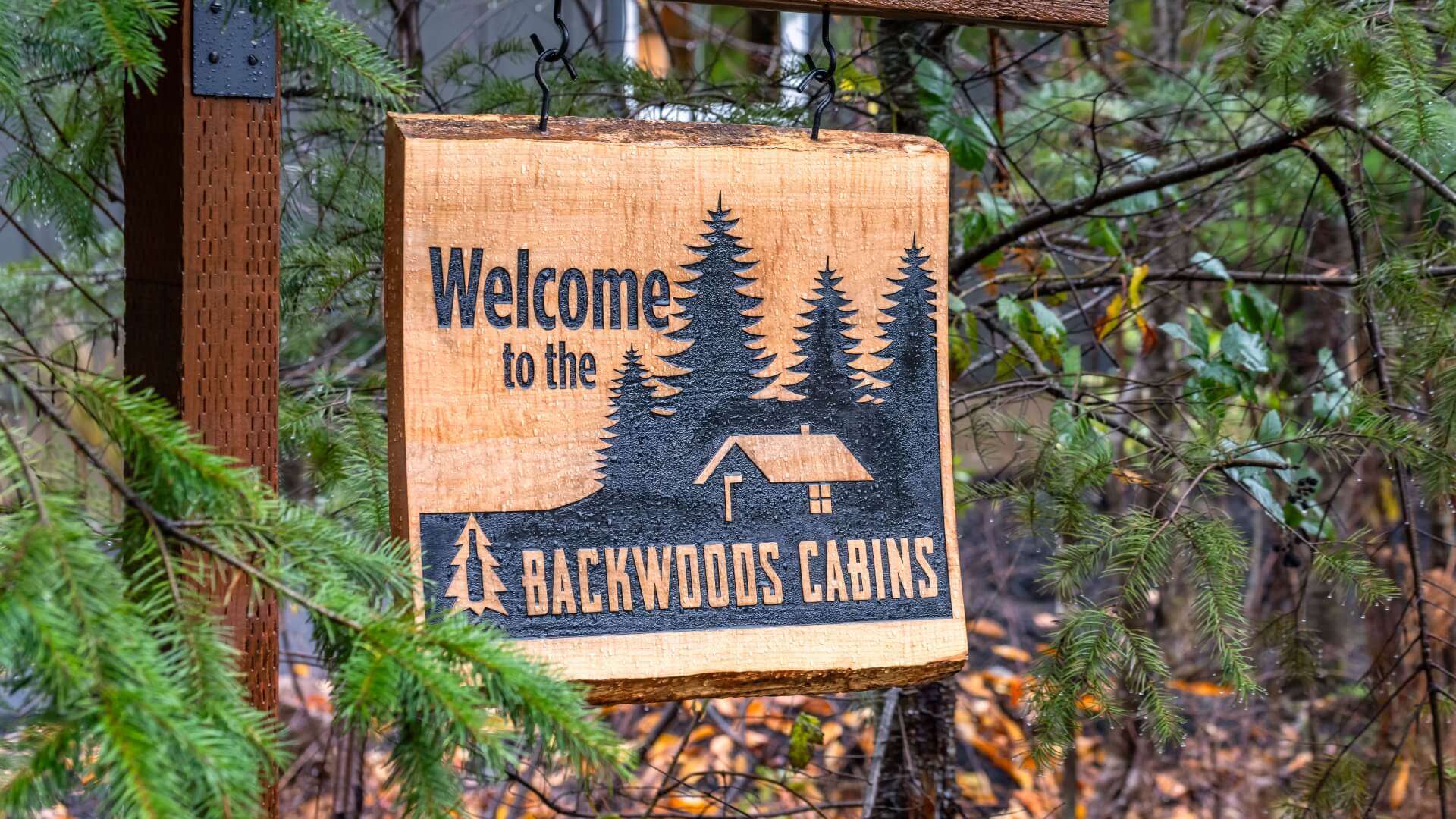 Wooden sign reading "Welcome to the Backwoods Cabins" surrounded by trees.