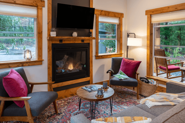 Cozy cabin sitting area with warm wood walls, a glowing fireplace, comfy chairs, and big windows looking out to the trees.
