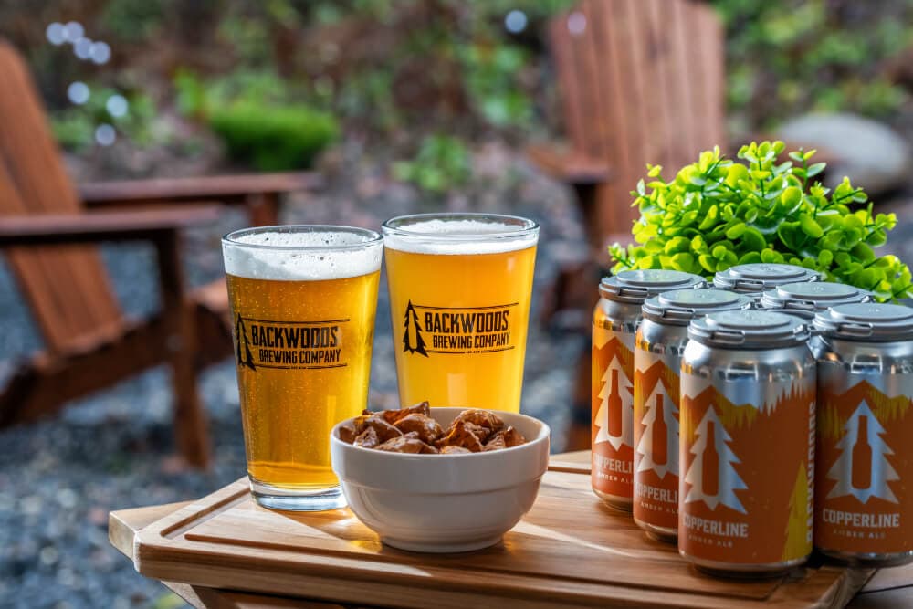 Two glasses of beer and a bowl of snacks on a wooden tray, with a six-pack of beer cans and greenery in the background.
