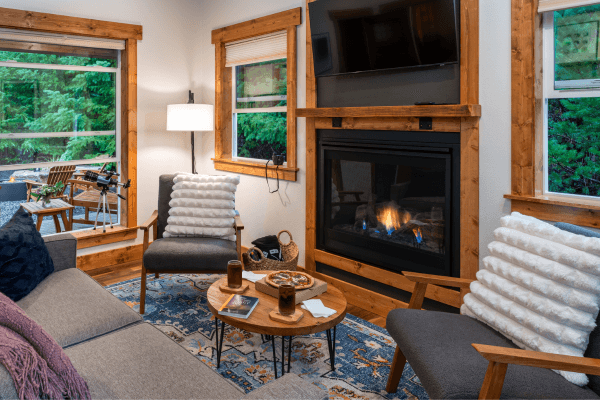Cozy cabin sitting area with warm wood walls, a glowing fireplace, comfy chairs, and big windows looking out to the trees.