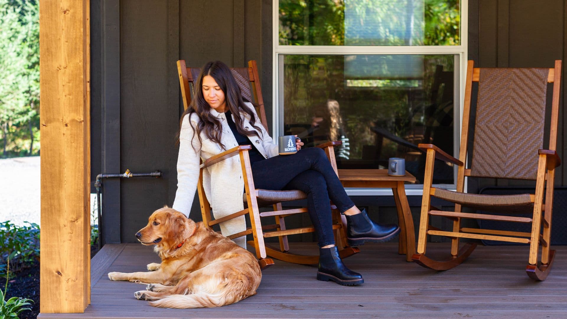 A woman sits on a porch, petting a golden retriever, with rocking chairs and coffee mugs nearby.