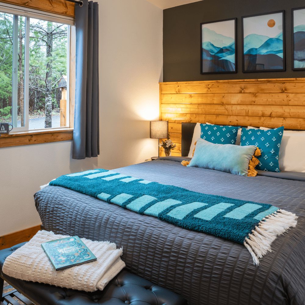 Image of king bed with luxurious grey comforter and turquoise pillows. View out window to Cascade Forest and mountain paintings above headboard.