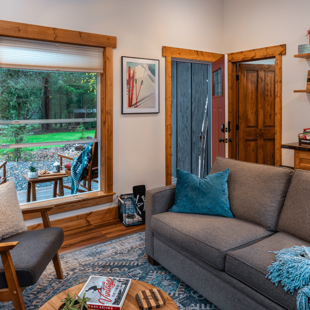 Image of cabin accommodations with view out to Cascade forest. Grey sofa and round table pictured with front door open.